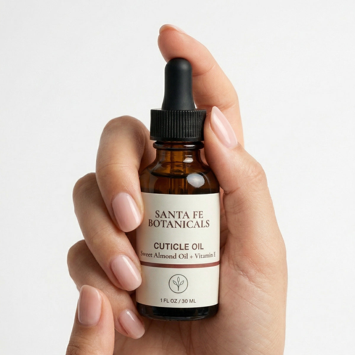 Manicured hand with natural pink nails holding Santa Fe Botanicals cuticle oil amber bottle