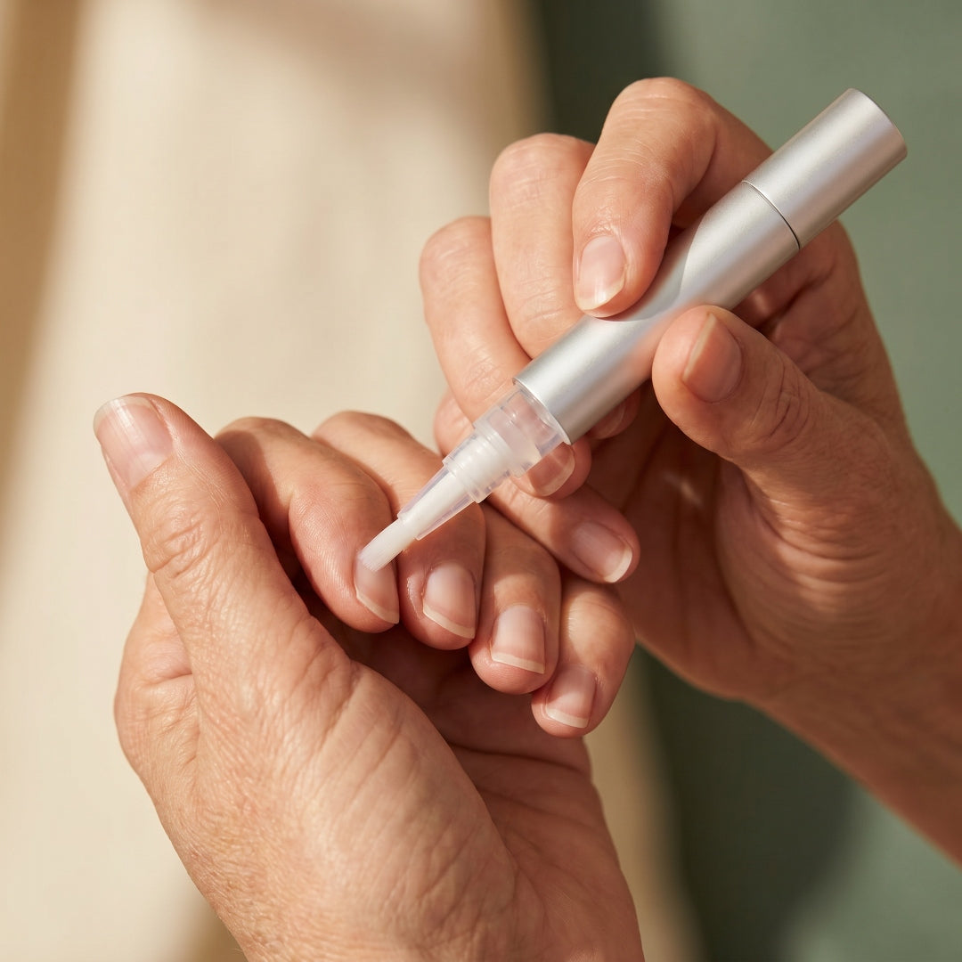 Fine nylon brush applicator of the Botanic Cuticle Serum Travel pen pressed to a cuticle against a sage-green backdrop
